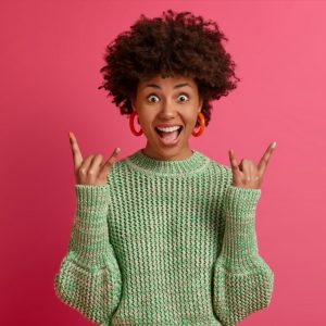 happy carefree dark skinned rebellious young woman enjoys awesome music makes rock n roll gesture has fun on music festival or cool event wears casual jumper poses against pink wall 1